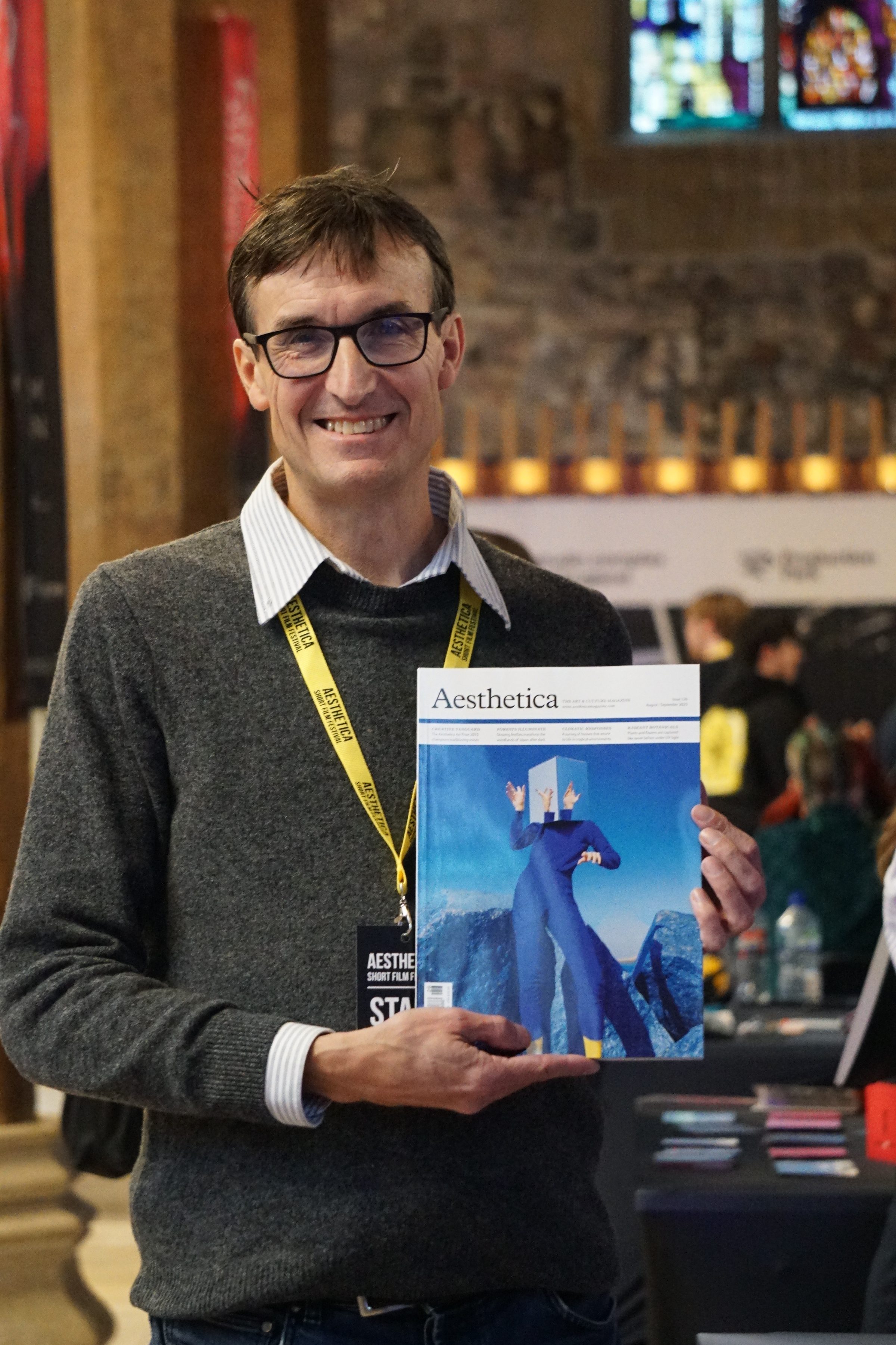 Dale Donley holding up one of his magazines, Aesthetica. He is smiling into the camera, quite pleased with himself. He's achieved a lot, so it's well deserved. His hair is short, with glasses on his face. He wears a grey jumper with a smart collar underneath. 