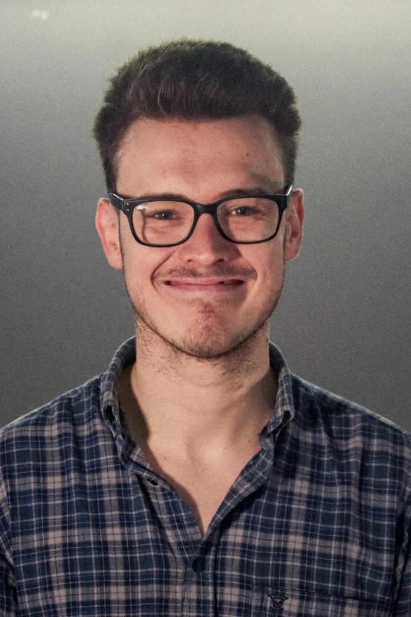 James Learoyd looks into the camera grinning with glasses, with short, brown hair pointing up. He wears a flannel shirt, and looks a bit like a teddy bear because he is one. 