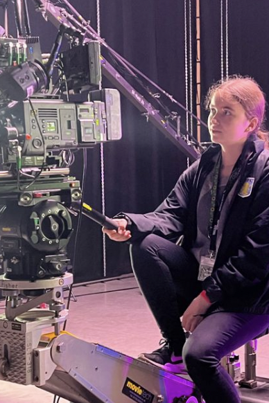 Rachel Radford sitting on a very expensive looking setup, which allows her to control an even more expensive camera. The room is lit up by a pink light, which reflects on her smiling face. 