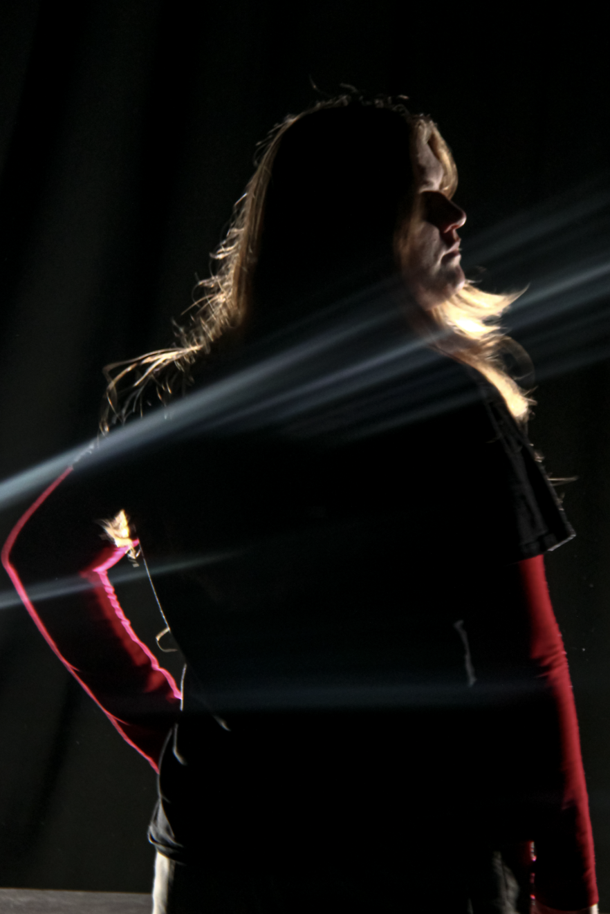 Zoe Seaton looking very super-hero like as she glances to her side, with an arm on her hip. The photo is in a dark room, with only her outline illuminated. 